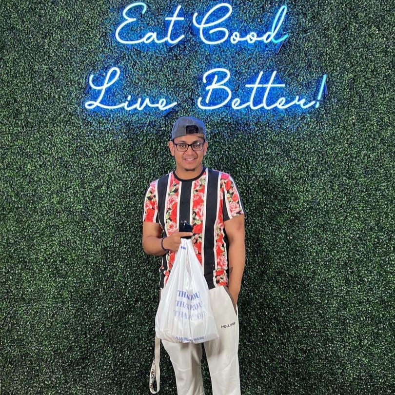 Man holding a Supreme Empanadas takeout bag in front of a green wall with a neon sign.