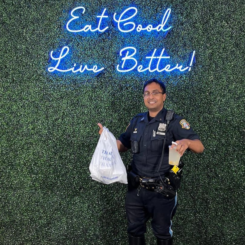 A police officer holding a Supreme Empanadas takeout bag and drink, standing before a neon sign.
