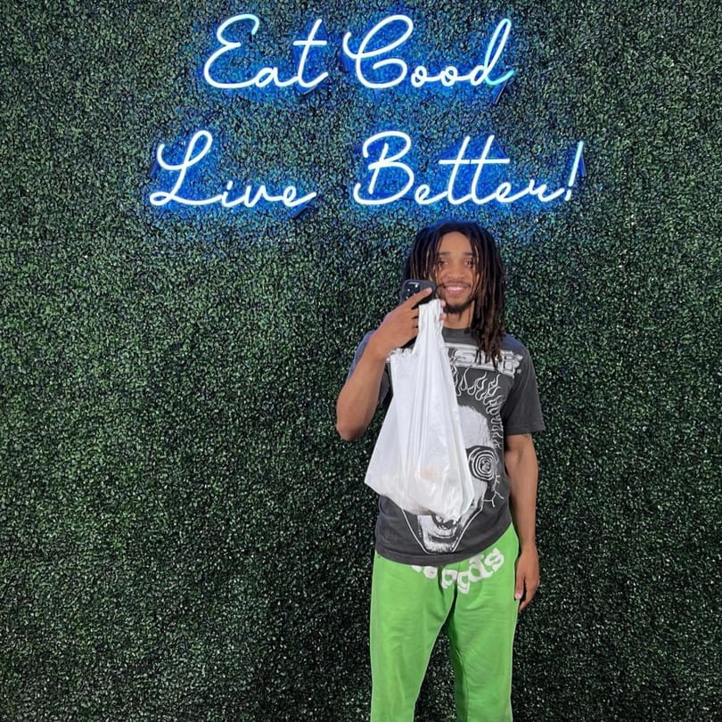 A man holding a white bag stands in front of a green wall with a blue neon sign.