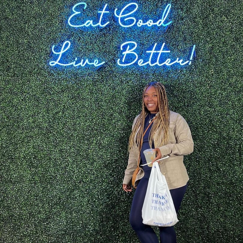 A woman smiles, holding a drink and a Supreme Empanadas takeout bag in front of a neon sign.