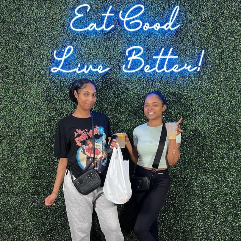 Two women holding drinks and a takeout bag, standing in front of a green wall with a neon sign.
