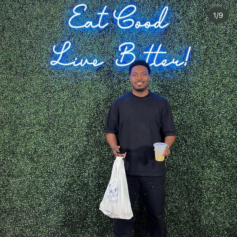 A man holding a Supreme Empanadas takeout bag and drink in front of a green wall with a neon sign.