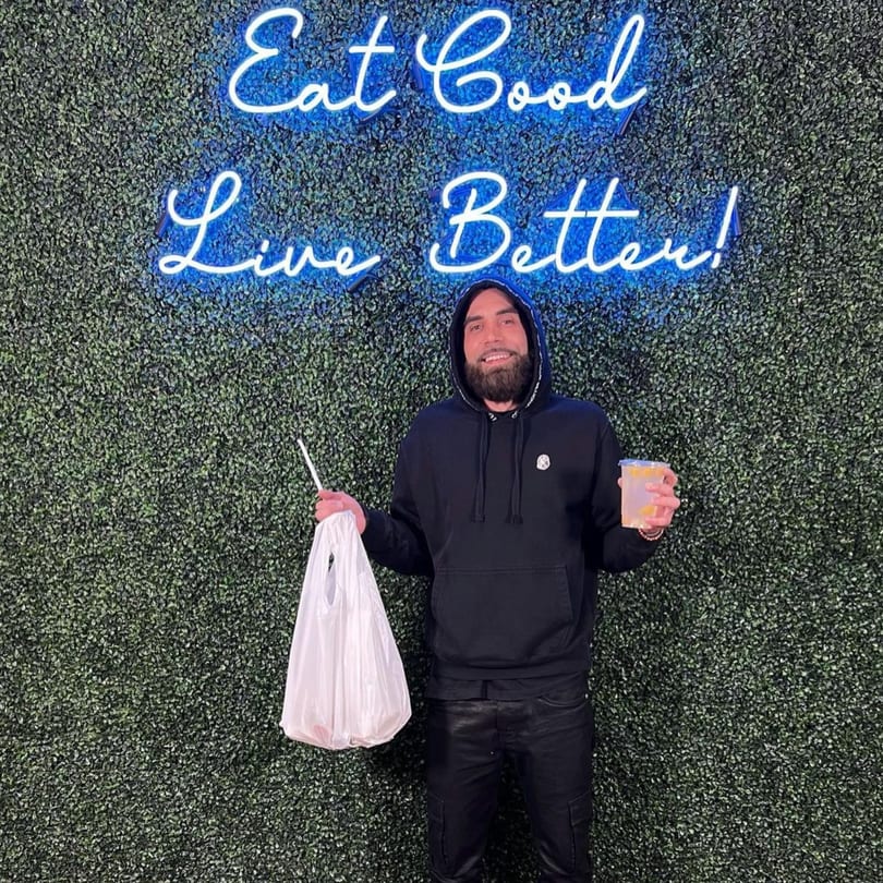 Man holding a Supreme Empanadas takeout bag and drink in front of a neon sign.