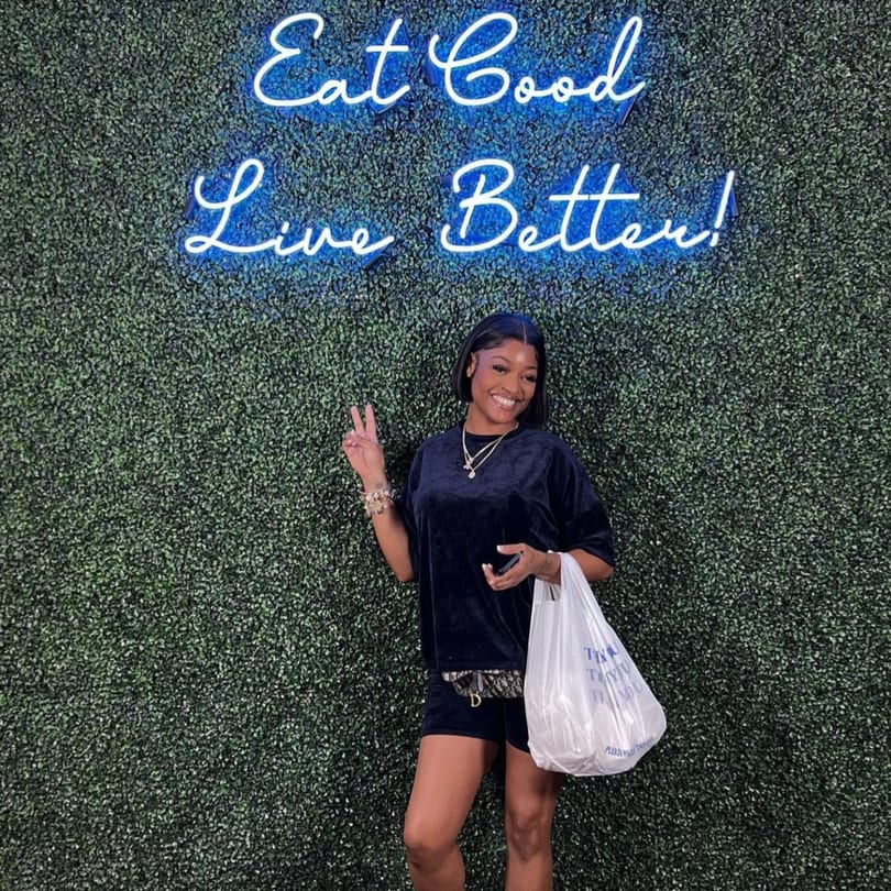 Smiling woman holding a Supreme Empanadas bag, posing in front of a green wall with a neon sign.