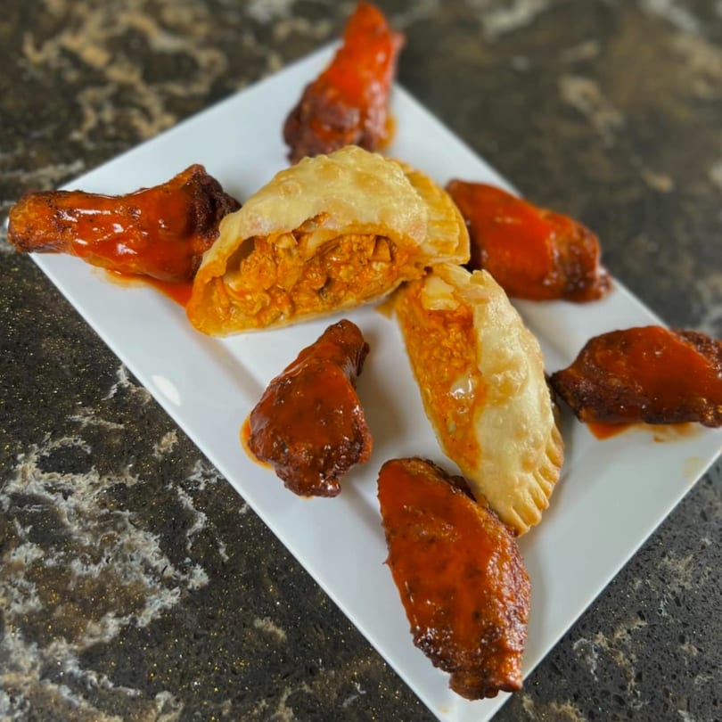 A white rectangular plate holds two Supreme Empanadas and several buffalo wings with red sauce.