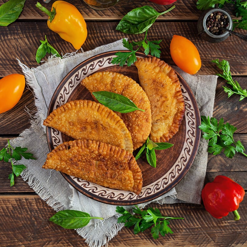 Four golden-brown Supreme Empanadas on a decorative plate, garnished with basil and parsley, surrounded by peppers.
