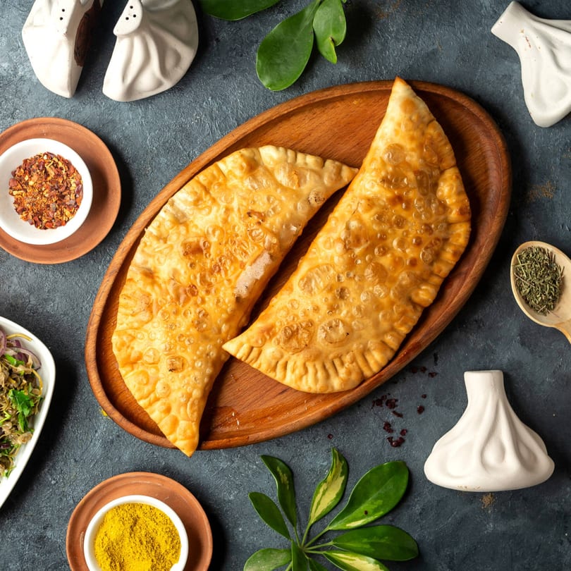 Two golden-brown Supreme Empanadas on a wooden tray, surrounded by spices and green leaves.